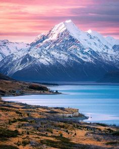 the mountains are covered in snow and there is a lake below them with grass on both sides