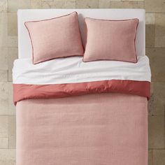 a pink and white bed with two pillows on top of the headboard in front of a beige brick wall