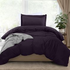 a bed with purple comforter and pillows in front of a window, next to a potted plant