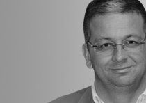 a black and white photo of a man with glasses smiling at the camera, in front of a gray background