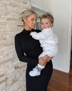 a woman holding a baby in her arms while standing next to a white brick wall