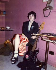 a woman sitting on the floor with a guitar in front of her and a pink wall behind her