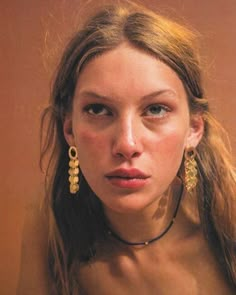 a woman with long hair and earrings on her neck looking at the camera while wearing gold jewelry