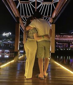 two people standing on a dock at night with their arms around each other, embracing