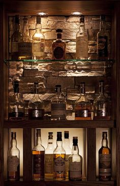 an assortment of liquor bottles on display in a wooden shelf with lights above them and stone wall behind it