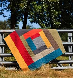 a colorful quilt sitting on top of a wooden bench next to a tree and fence