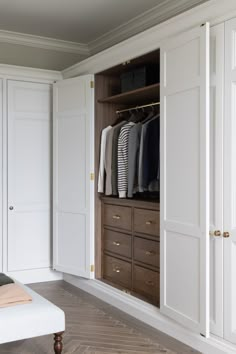 an open closet with clothes hanging on the doors and drawers in front of it, next to a white bench