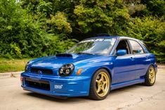 a blue subarunt is parked in front of some trees and bushes with gold rims