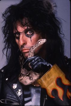 a man with long black hair and makeup holding a snake in his right hand while wearing a leather jacket