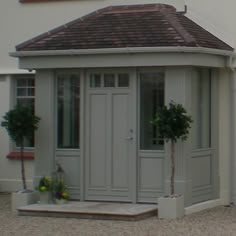 a small white building with a brown roof and two potted trees on the side