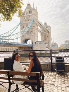 two young women sitting on a bench in front of the tower bridge, london england