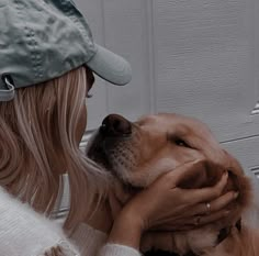 a woman is petting her dog in front of a white wall and wearing a baseball cap