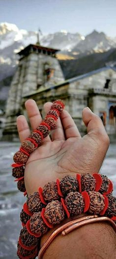 a hand holding some kind of bracelet in front of a building with snow covered mountains behind it