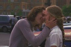 two people kissing each other in front of a car on the street with cars behind them