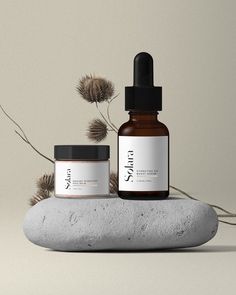 two bottles of skin care sitting on top of a rock next to some dry grass