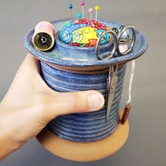 a hand holding a blue container with some pins and scissors in it, on top of a table