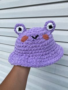 a purple crocheted hat with eyes and nose is held up in front of a white wall