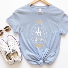 Embrace your Catholic faith with our Miraculous Medal T-Shirt. Miraculous Medal holds additional symbolism and imagery. It typically features a cross intertwined with the letter "M," representing the intersection of Jesus Christ's sacrifice and the Virgin Mary's role in salvation. This combination signifies the inseparable connection between Jesus and His mother, highlighting Mary's pivotal role in the redemption of humanity. Surrounding the cross and "M," there are twelve stars, representing th Catholic Outfits, Catholic T Shirts, Catholic Tshirt, Catholic Shirts, Catholic Clothing, Catholic Shirt, Catholic Tshirts, Catholic Decor, Cross Shirt