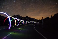 the night sky is filled with stars and colorful light trails on an empty road in front of mountains