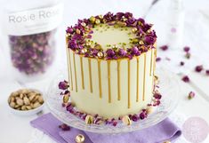 a white cake with purple flowers on top and gold decorations around the edges, sitting on a glass platter