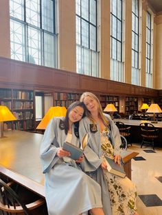 two women in robes are sitting on a bench and posing for the camera with bookshelves in the background