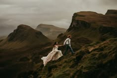 a bride and groom walking up the side of a mountain holding each other's hands