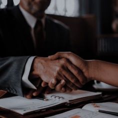 two people shaking hands while sitting at a table in front of paperwork and papers on top of each other