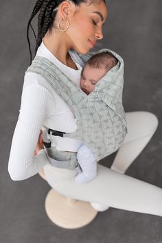 a woman holding a baby in a grey wrap on top of a white chair and looking down at it