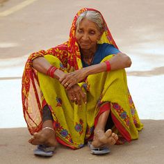 an old woman sitting on the ground with her feet crossed and wearing a yellow sari