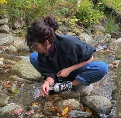 a woman crouches down to collect water from a stream with rocks and leaves on the ground