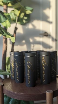 four black cups with gold lettering on them sitting on a table next to a potted plant