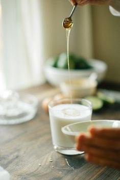 a person pouring some liquid into a glass on top of a wooden table with cucumbers in the background