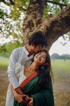 a man and woman standing next to each other in front of a tree with their arms around each other