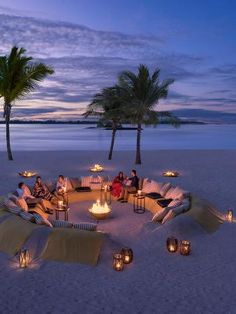 people sitting around a fire pit on the beach at dusk with palm trees in the background