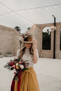 a woman wearing a hat and holding a bouquet in front of a brick wall with string lights