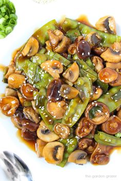 a white plate topped with green beans and mushrooms covered in sauce next to spoons