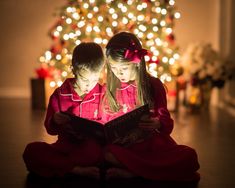 two young children sitting on the floor reading a book in front of a christmas tree