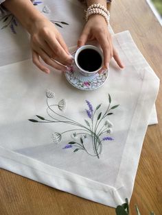 a woman holding a coffee cup on top of a white table cloth with purple flowers