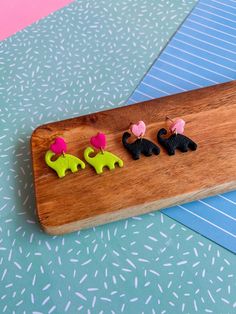 three small elephant shaped earrings are on a wooden board next to blue and pink paper