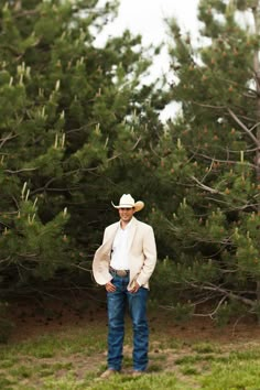 a man wearing a cowboy hat standing in front of some trees and grass with his hands in his pockets