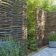 a wooden fence made out of bamboo sticks and plants next to a sidewalk with blue flowers in the foreground