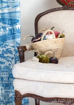 a wicker basket filled with fruit sitting on top of a white chair next to a blue and white curtain