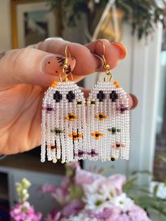 a person holding up two pairs of beaded owl earrings in their left hand, with flowers behind them