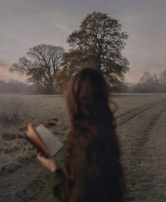 a woman standing in the middle of a field holding an open book and looking at trees