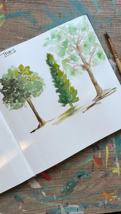 an open book with watercolor trees on it next to a pencil and paintbrush