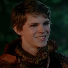 a young man smiling at the camera in front of some green trees and bushes, wearing a scarf