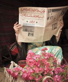 a woman reading a newspaper while sitting on a chair with flowers in front of her