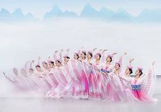 a group of women dressed in pink and white dance on stage with mountains in the background