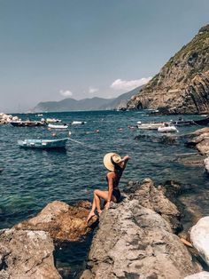 a woman in a hat sitting on rocks near the water with boats floating around her