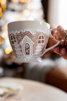 a person holding a cup with a gingerbread house design on it and a christmas tree in the background